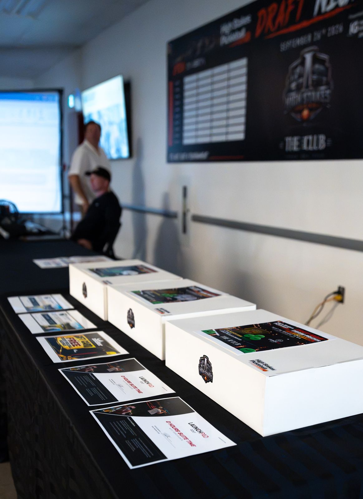 A black-draped table lined with white prize boxes and printed certificates, with the Draft Night board on the wall behind and two people out of focus in the background.