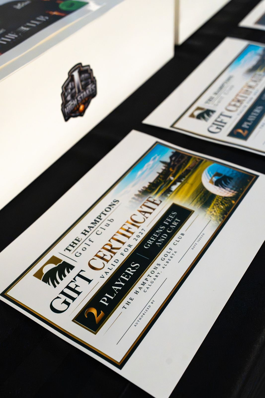 Close-up of a golf course gift certificate lying on a black table beside a white prize box carrying the High Stakes Series sticker.