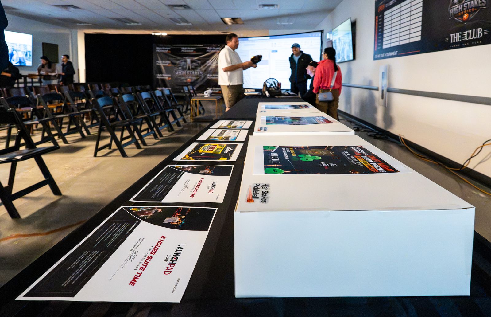 Wide low view along a table of white prize boxes and certificates toward a room with chairs, a High Stakes banner and a few people talking.