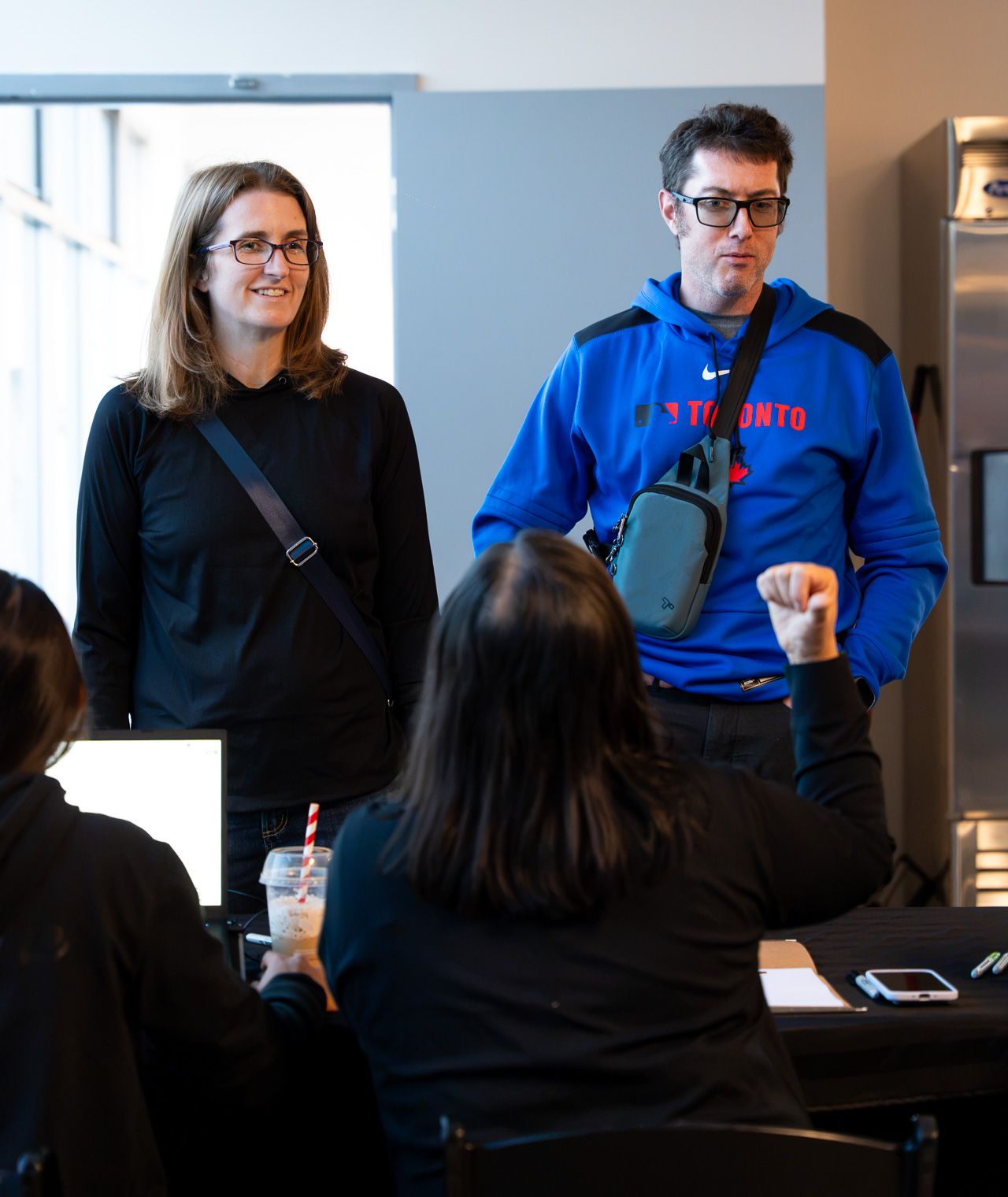 Two guests smile at the check-in table while a volunteer in the foreground gestures at them.