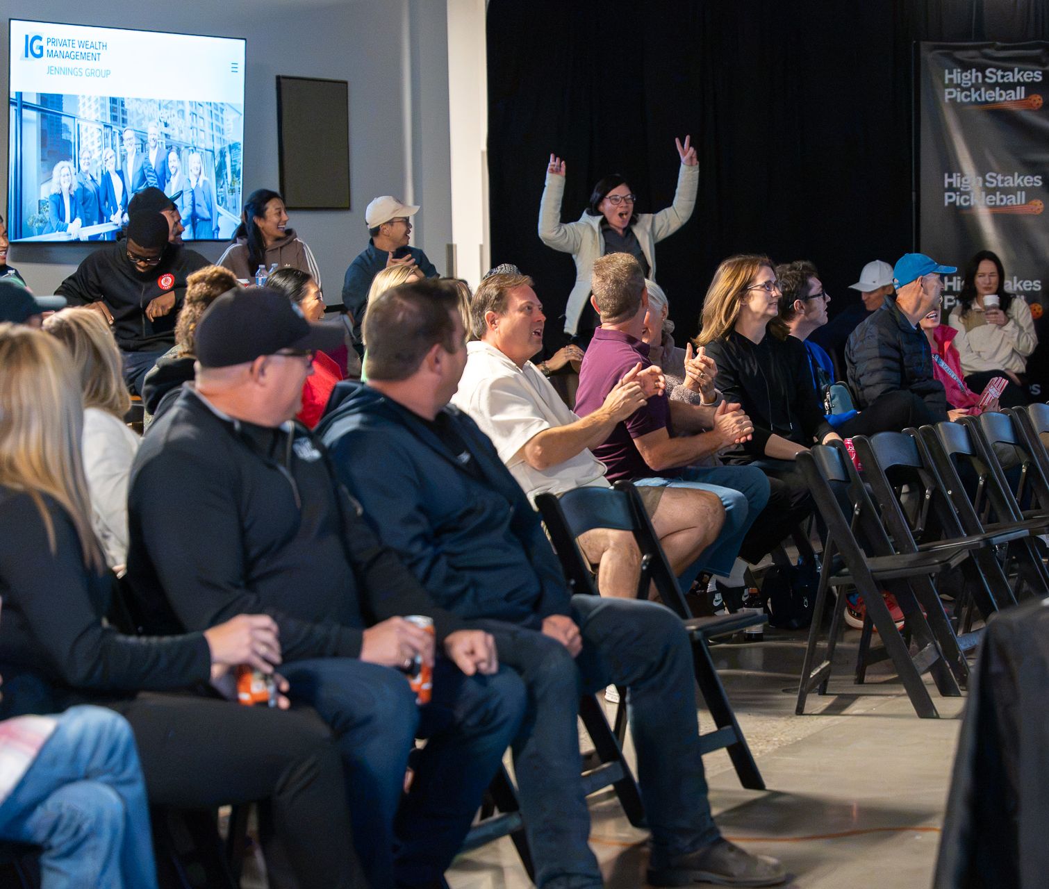 A guest raises both arms and cheers behind rows of seated spectators, with an IG Private Wealth Management slide on a TV and a High Stakes Pickleball banner at the right.