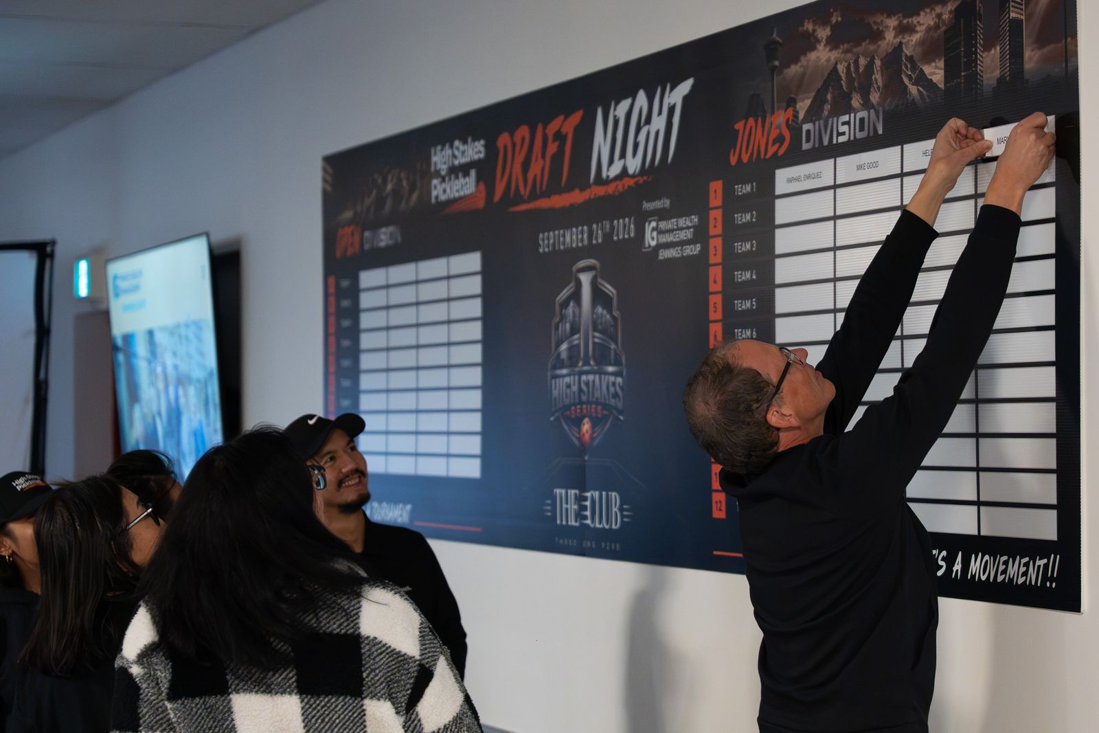 A man in black reaches up to slot a name card into the Jones Division column of the Draft Night board while three people watch.