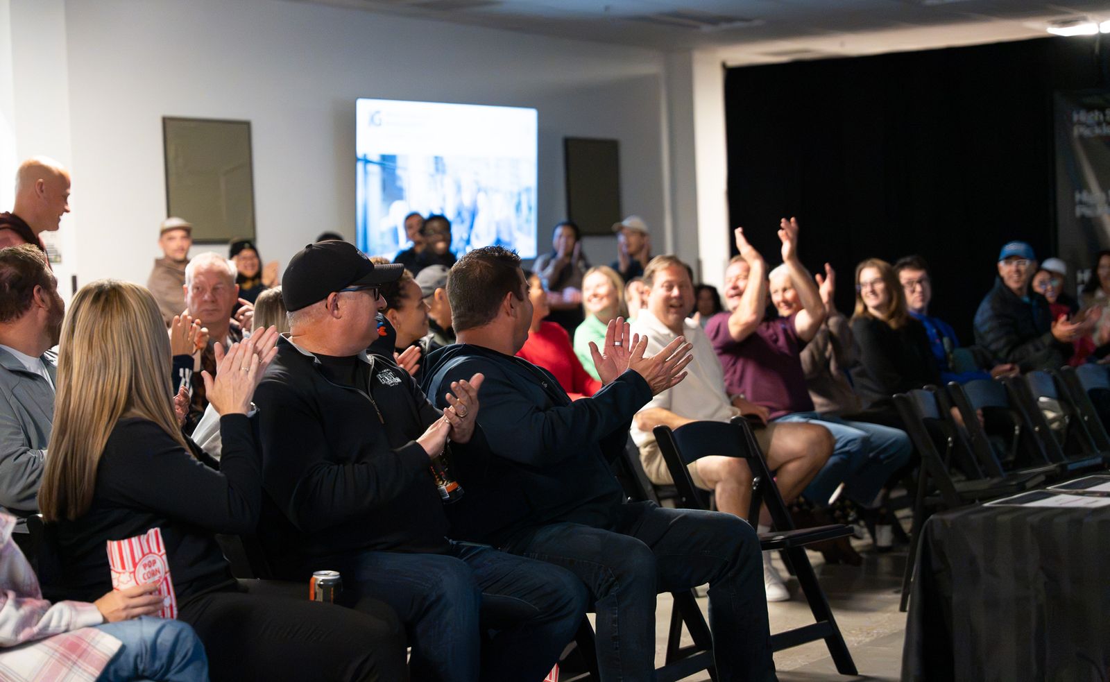 Seated spectators clap and cheer, one man raising both arms, in a room with an IG slide on the TV and a black curtain at right.