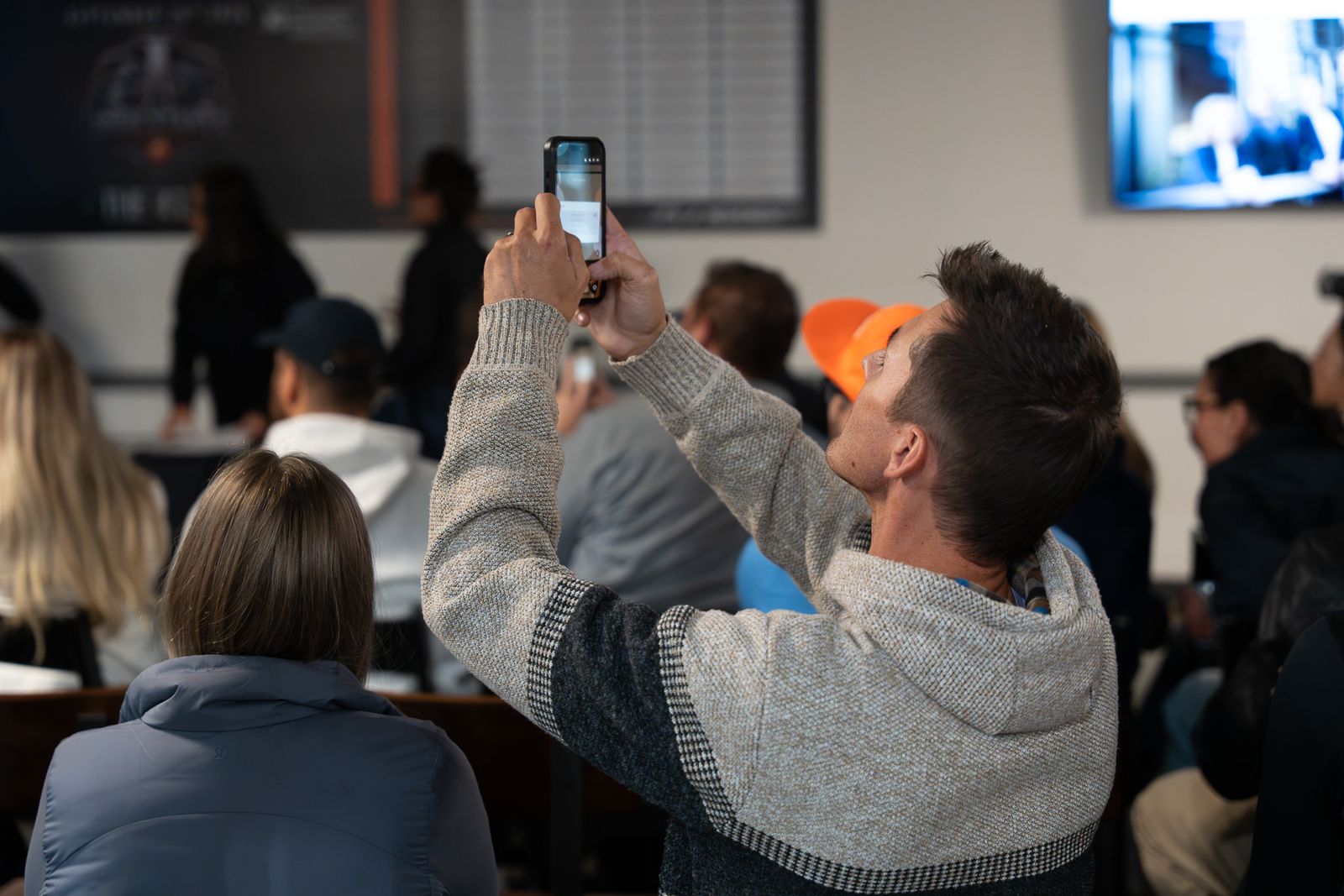 A man in a patterned knit hoodie holds his phone up to take a photo during the presentation, with seated guests and a High Stakes board behind him.