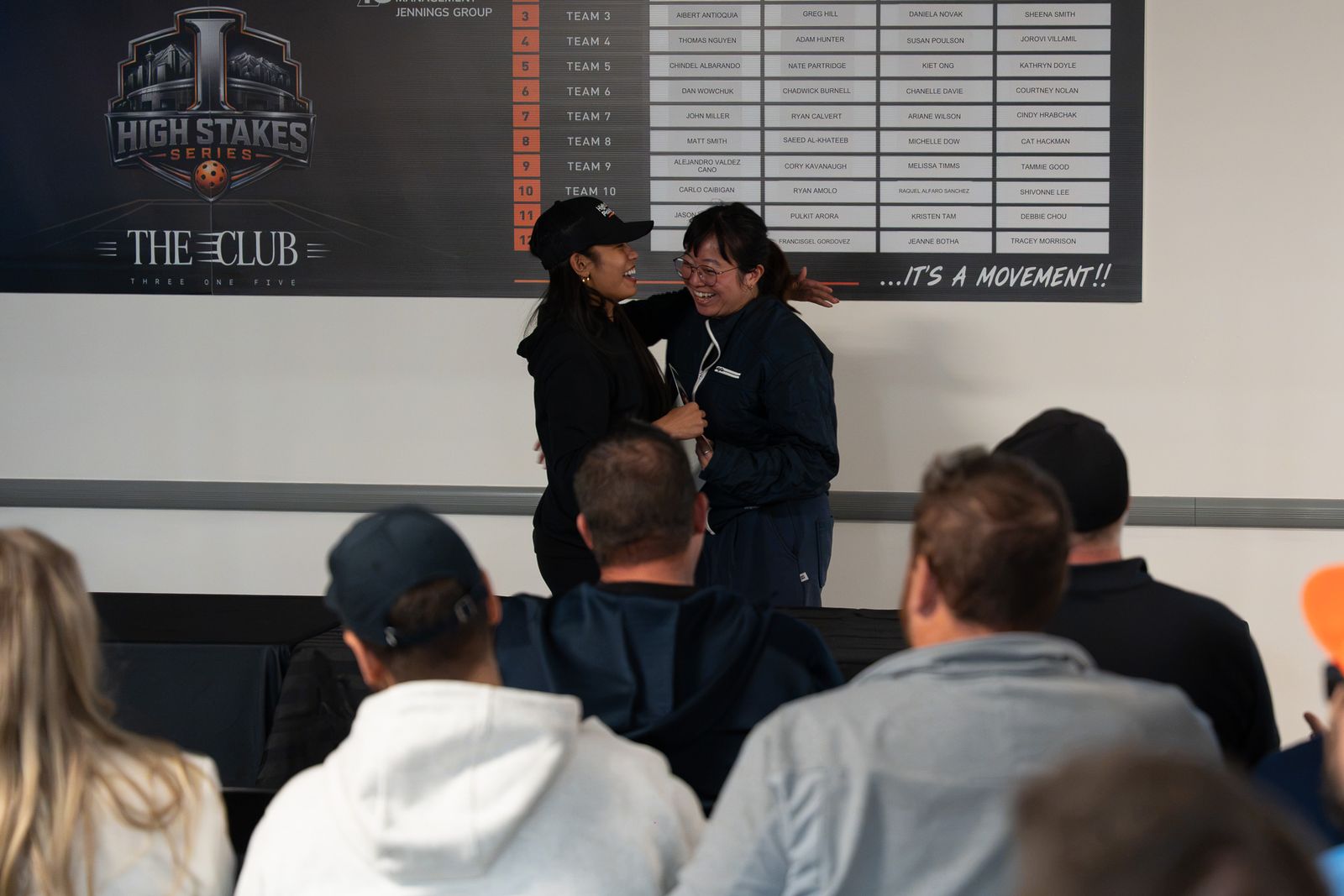 Two women embrace and laugh in front of the High Stakes Series Draft Night board while seated guests watch from the foreground.