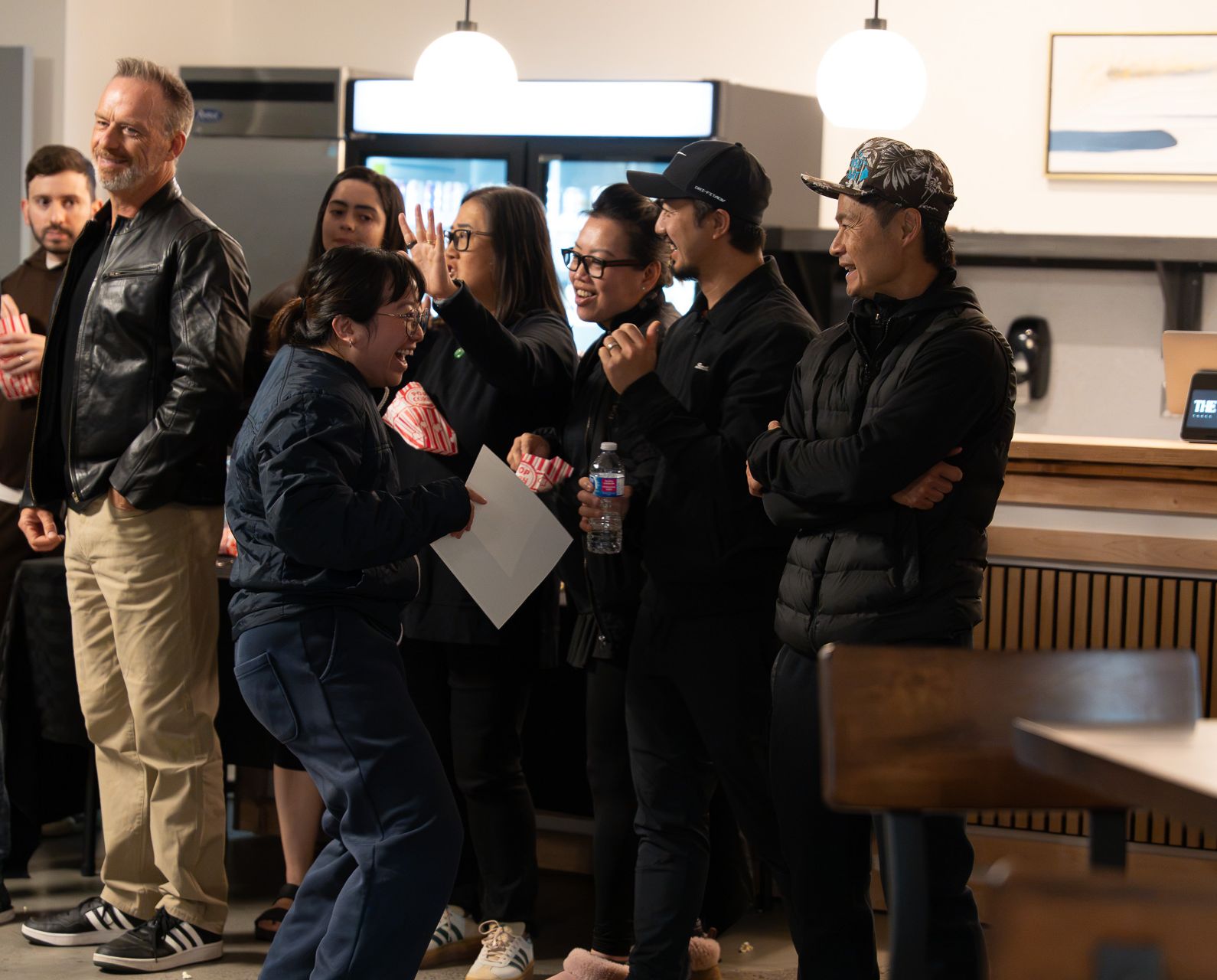 A group of guests laugh and react together near a refreshment area, one woman bending forward with a sheet of paper and popcorn boxes around.