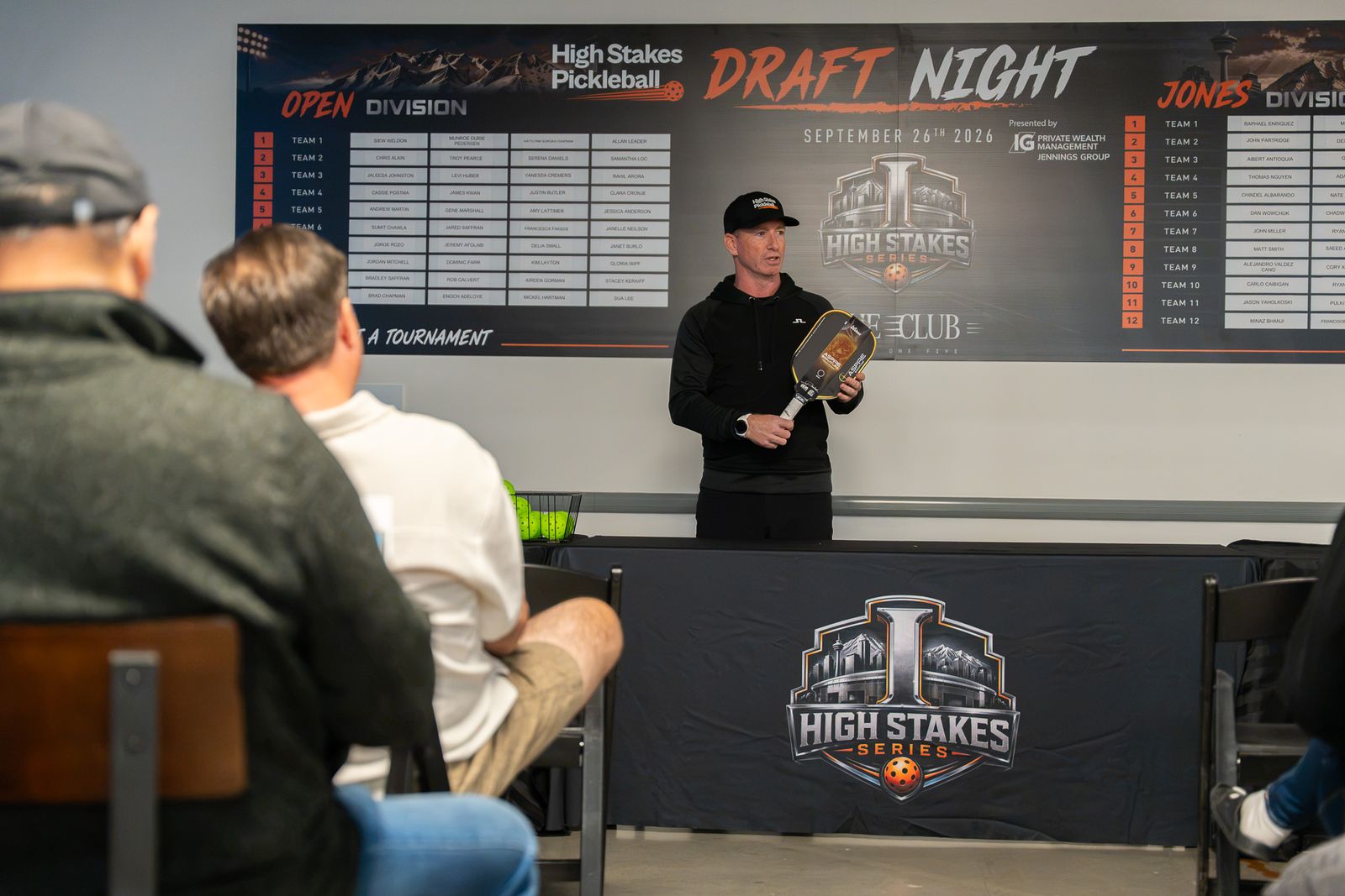 The founder in a black High Stakes cap and black hoodie holds up a pickleball paddle while speaking, in front of the Draft Night board and High Stakes Series banner.